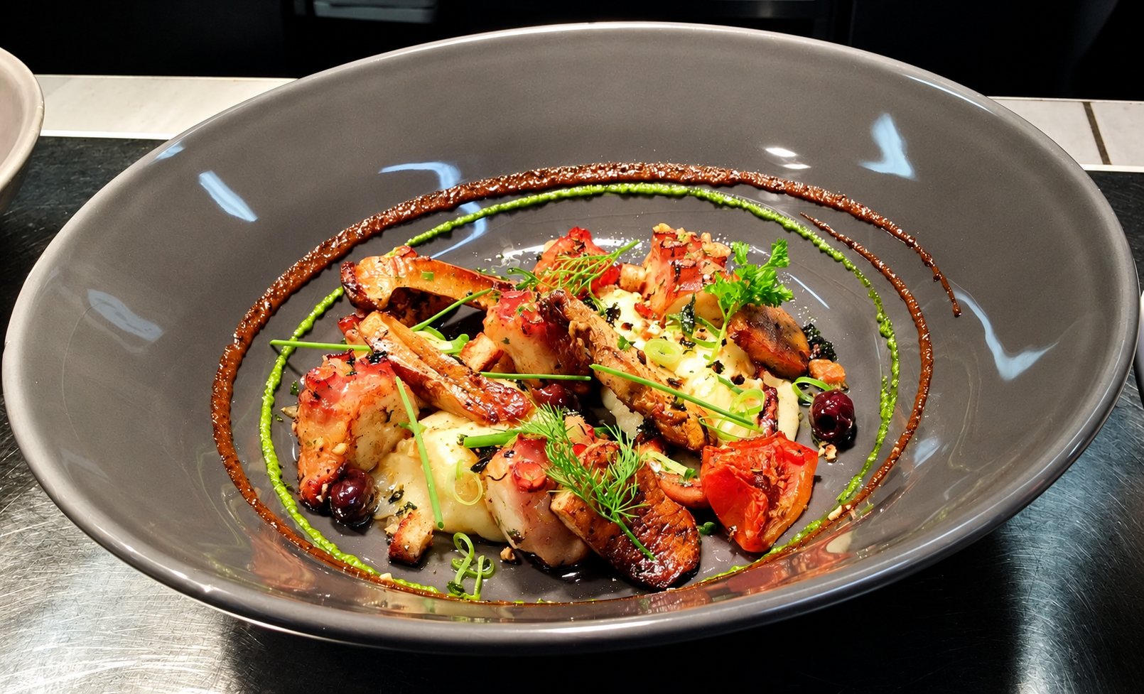 Un plat raffiné de viandes grillées, tomates et herbes fraîches, nappé de sauces verte et brune, servi dans un grand bol gris posé sur un comptoir en inox.