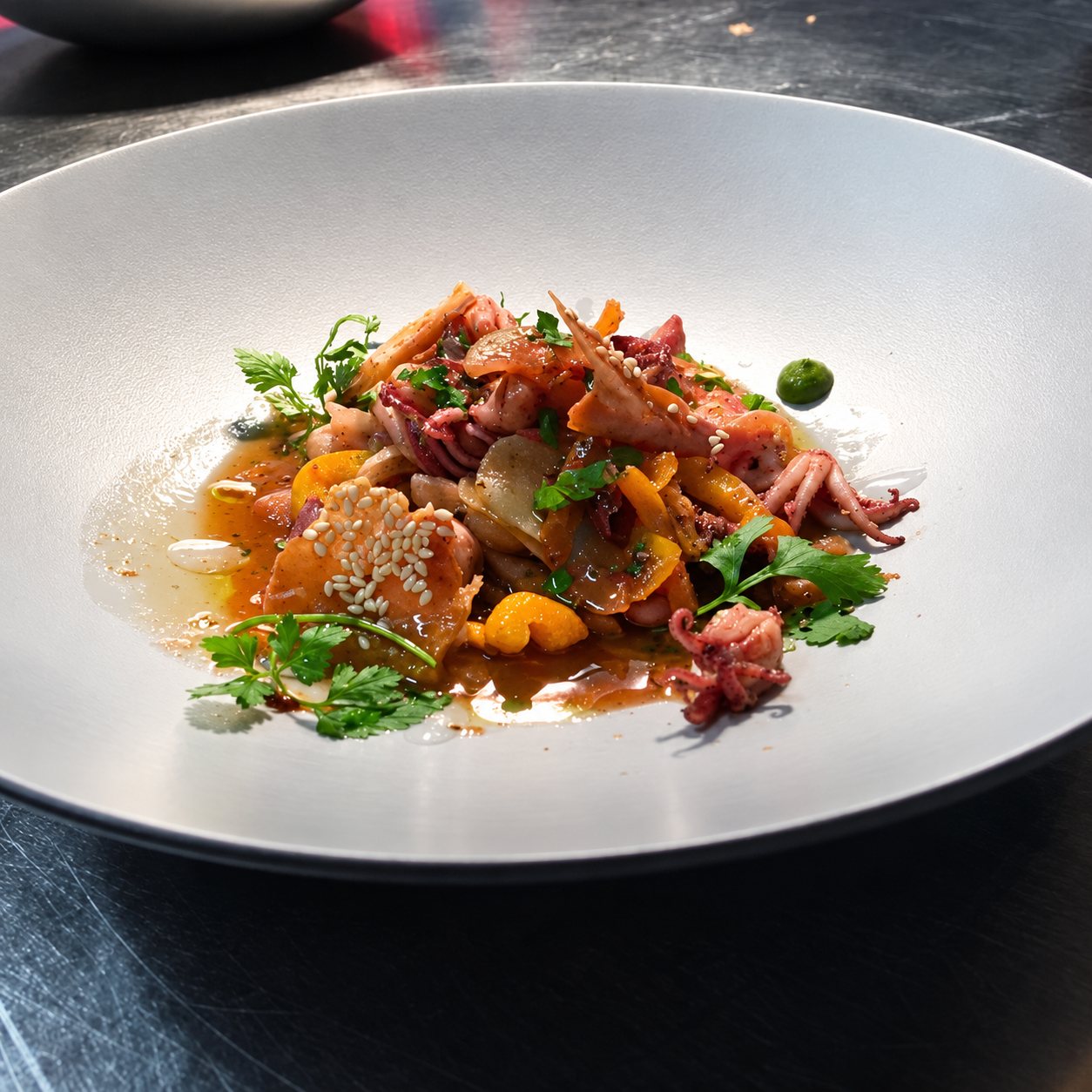 Une assiette creuse grise contient un plat dressé de calamars, de poivrons jaunes, de sésame et de jeunes pousses d'herbes dans une sauce brune.