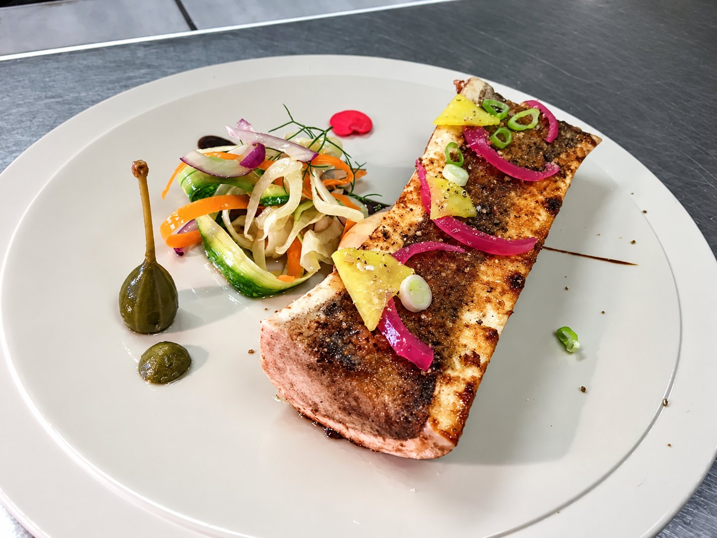 Un pavé de poisson grillé garni d'oignons rouges marinés, de ciboule et de citron confit, servi avec une salade de légumes en rubans et une câpre sur une assiette beige.