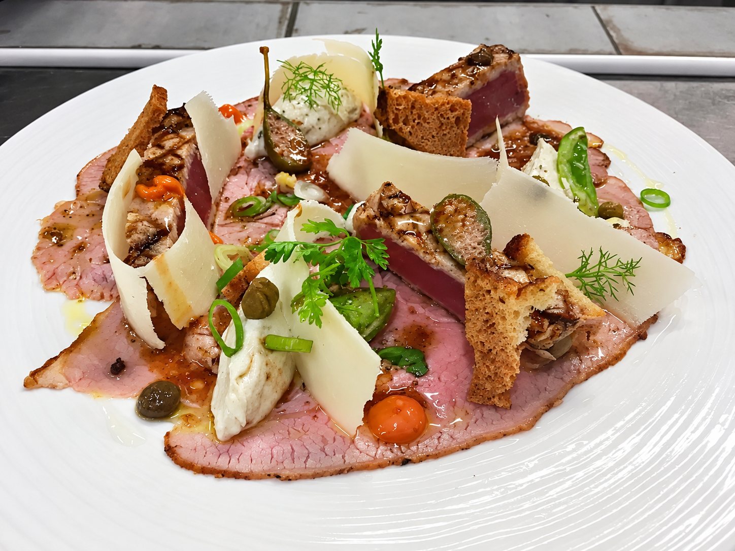 Une assiette blanche garnie de fines tranches de viande rosée, de morceaux de thon mi-cuit, de copeaux de fromage, de croûtons, de câpres et d'herbes fraîches.