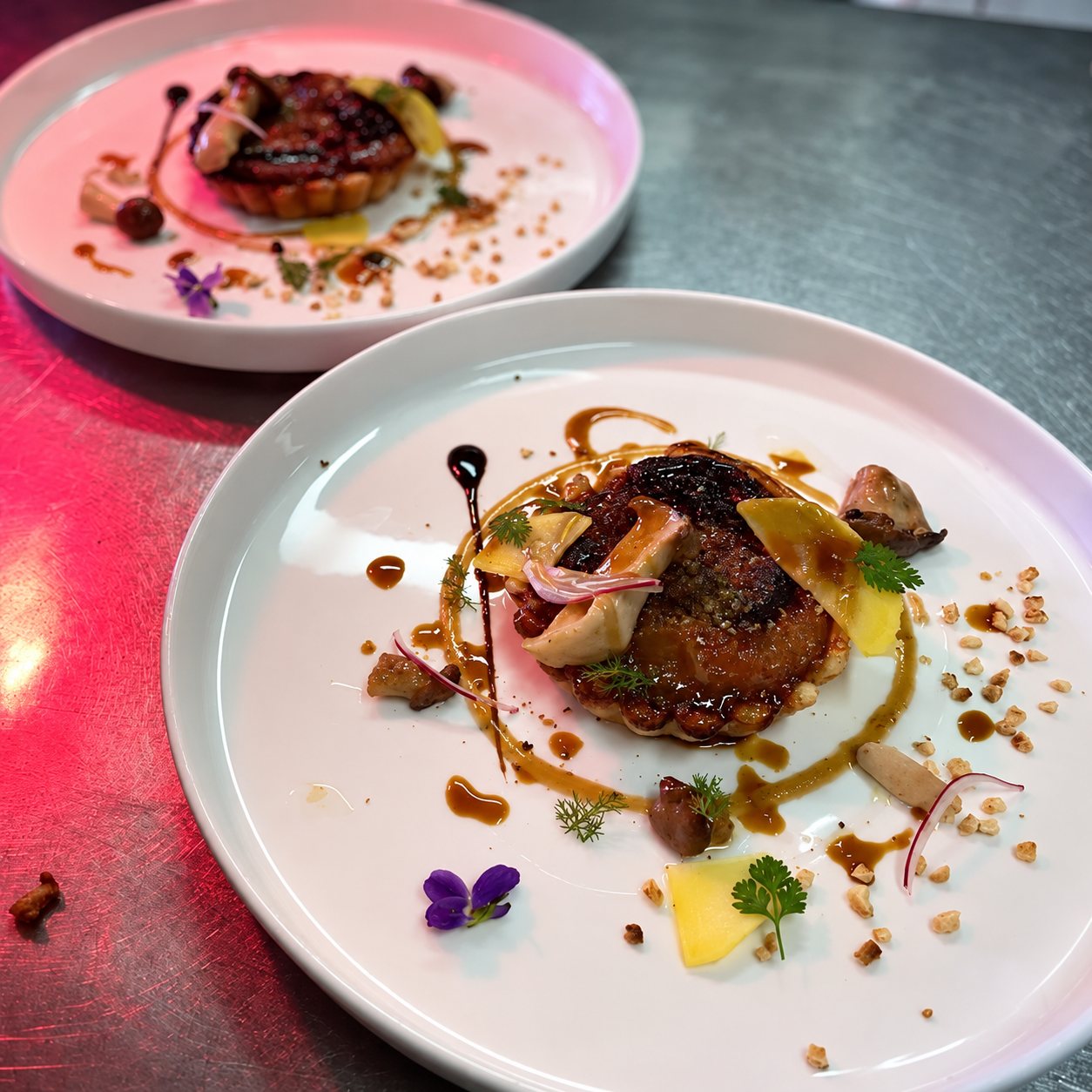 Deux assiettes blanches contenant chacune une tartelette dressée avec sauce, fleur de violette, herbes fraîches, oignon rouge et éclats de noisettes, posées sur un plan de travail en inox éclairé de rouge.