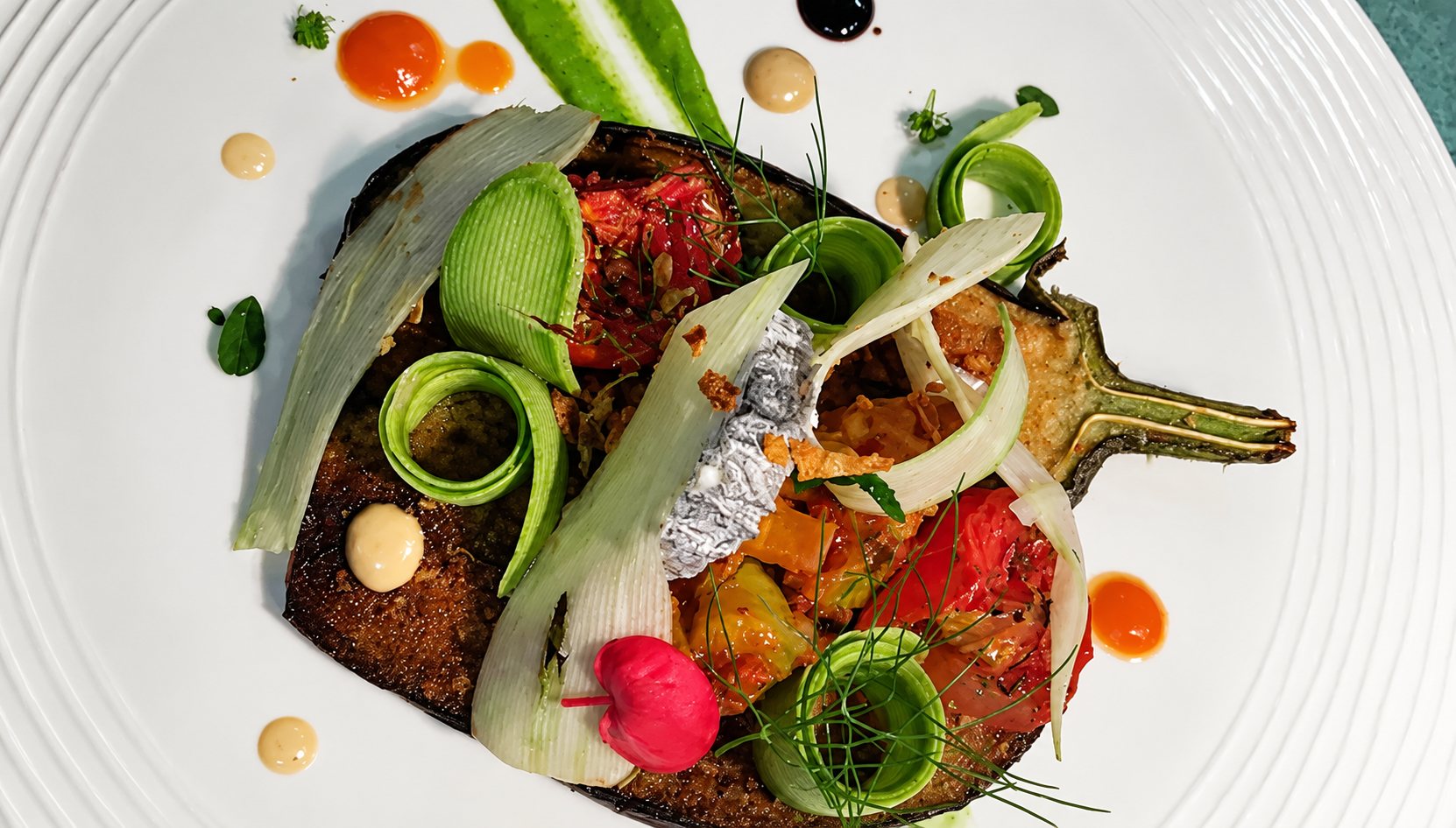 Vue de dessus d'une demi-aubergine rôtie dressée sur une assiette blanche, garnie de rubans de courgette, de fenouil, de tomates et de touches de sauces colorées.