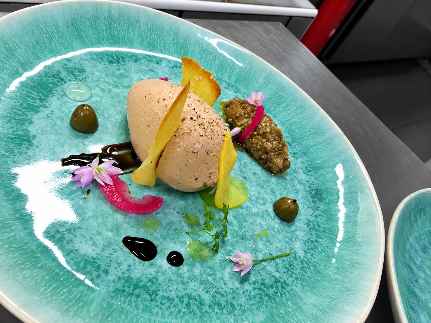 Assiette en céramique turquoise dressée avec une quenelle de mousse rosée, des chips jaunes, des câpres, des fleurs de ciboulette et des touches de sauce sombre.