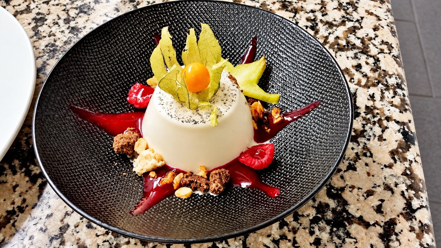 Une panna cotta à la vanille décorée d'un physalis, de coulis de fruits rouges et de crumble, servie dans une assiette creuse sombre posée sur un plan en granit.