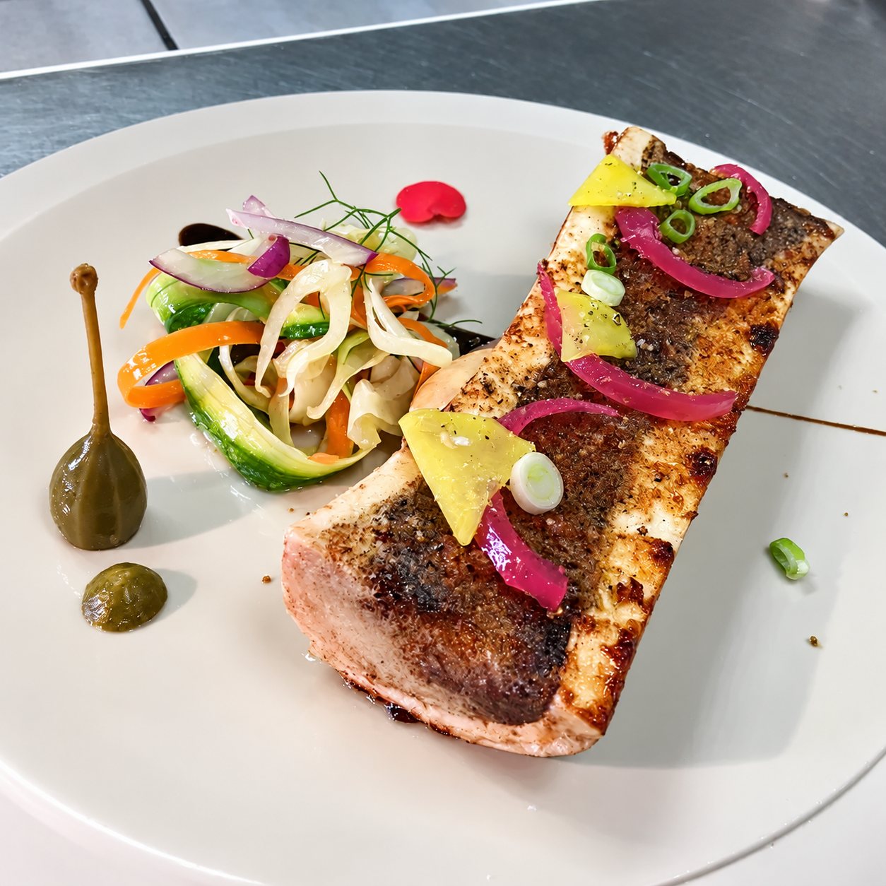 Une assiette beige présente un pavé de poisson grillé garni d'oignons rouges marinés et de ciboule, accompagné d'une salade de légumes en rubans, d'une câpre et d'une pointe de sauce verte.