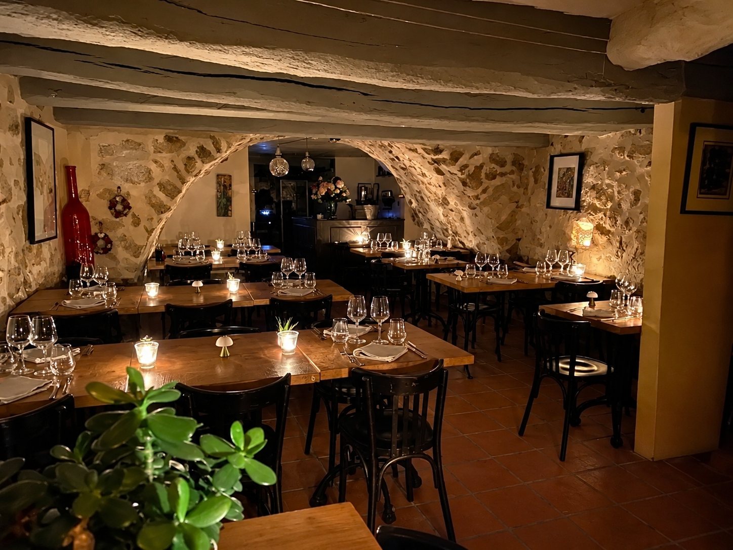 Salle de restaurant à l'ambiance tamisée avec des murs en pierre apparente, des poutres au plafond et des tables en bois dressées avec des verres et de petites bougies.