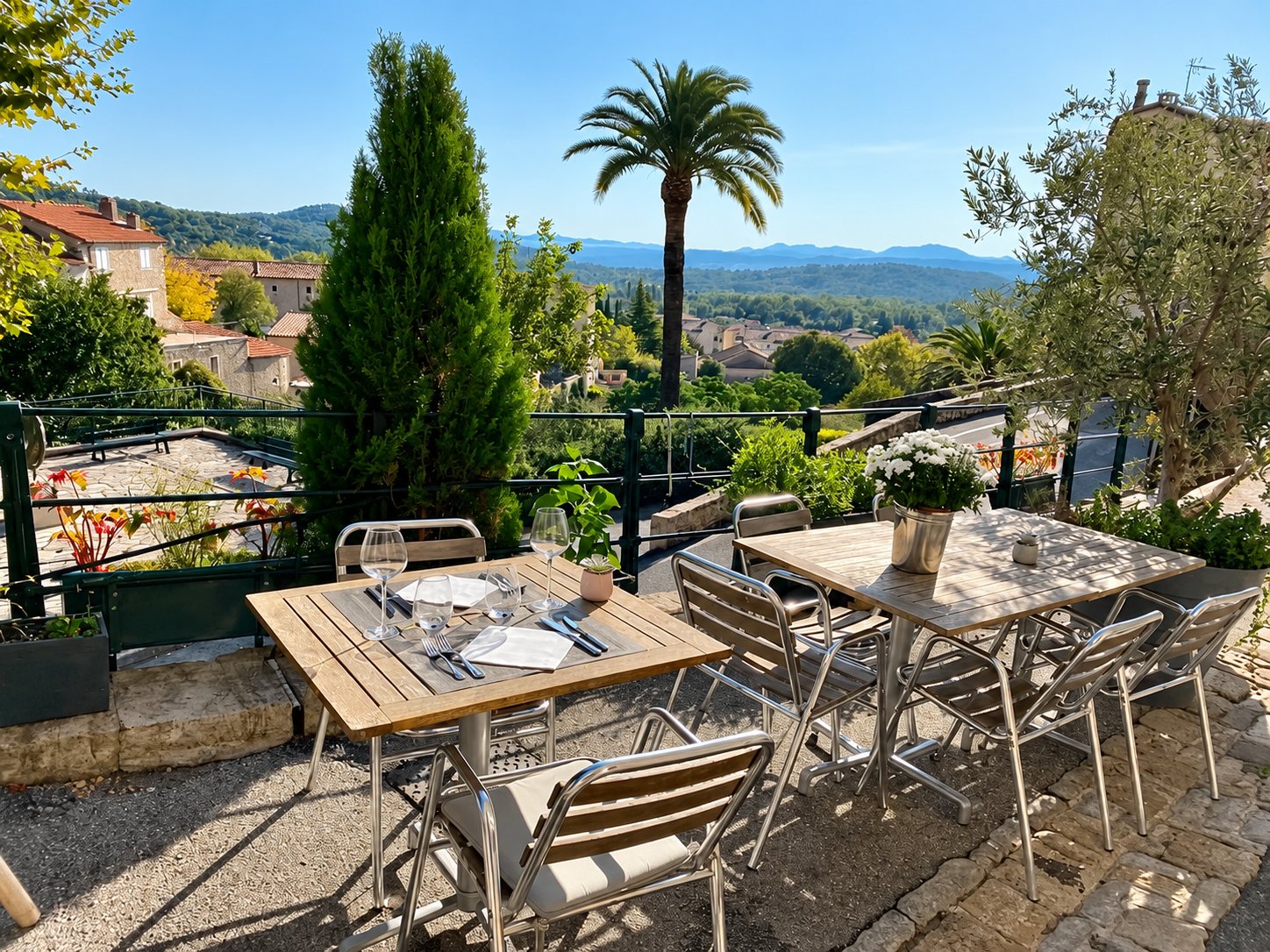 Des tables dressées en bois et des chaises en aluminium sur une terrasse ensoleillée, avec un palmier, des oliviers et un village provençal en contrebas.